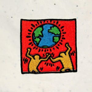 Könnte beinhalten: Gestickter Aufnäher mit rotem Hintergrund, blau-grüner Weltkugel und zwei gelben tanzenden Figuren. Der Aufnäher hat einen schwarzen Rand. Das Design erinnert an ein Kunstwerk von Keith Haring.