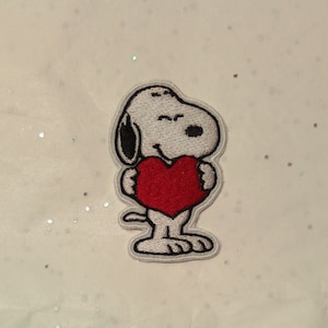 Può includere: Toppa ricamata con Snoopy dei Peanuts, che tiene un cuore rosso. La toppa ha uno sfondo bianco con un bordo argentato, adatta per abbigliamento o accessori.
