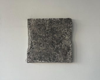 Può includere: Un'opera d'arte quadrata e strutturata, di colore grigio, con una superficie ruvida e irregolare. L'opera è montata su un muro bianco, creando un contrasto tra lo sfondo liscio e il pezzo d'arte strutturato. La tavolozza dei colori dell'opera è principalmente grigia.
