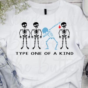 May include: White t-shirt with a graphic of four skeletons. One skeleton is dabbing, and another has a drop of blood above its head. The text "TYPE ONE OF A KIND" is printed below the skeletons. The shirt is on a light wood surface.
