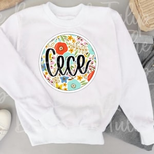 May include: White sweatshirt with a round floral design. The design features the name "Cece" in black script, surrounded by colorful flowers in red, blue, and yellow. The sweatshirt has a crew neck and long sleeves.