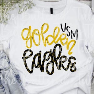 Könnte beinhalten: Weißes T-Shirt mit den Worten "Golden Eagles" in gelber und schwarzer Schrift. Das Shirt hat auch die Buchstaben "VSM" in Schwarz. Der Text ist mittig auf der Vorderseite des Shirts angeordnet.