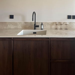 Pode incluir: Design moderno de cozinha com pia branca, torneira preta e armários de madeira escura. A bancada é bege claro com veios sutis. Uma luminária redonda com moldura preta e três taças de vinho completam a estética minimalista.