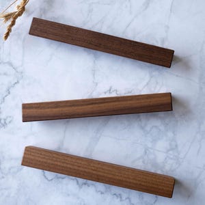 May include: Three rectangular, dark brown wooden bars arranged diagonally on a white marble surface. The bars have a smooth, polished finish. Dried wheat stalks and a white stone are in the background.