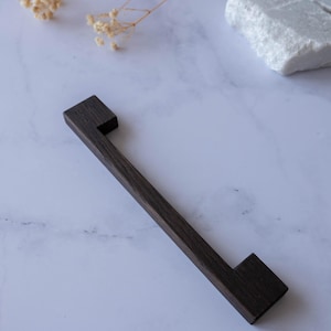 Könnte beinhalten: Ein dunkelbrauner Möbelgriff aus Holz mit einem linearen, rechteckigen Design und zwei quadratischen Befestigungsblöcken an jedem Ende. Der Griff hat eine glatte, matte Oberfläche und eine gerade, längliche Stange zwischen den Enden.