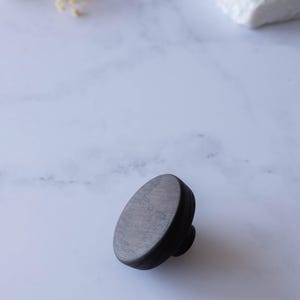 May include: A circular cabinet knob crafted from dark-toned wood with a visible grain pattern. The knob features a flat, rounded top surface and a smaller cylindrical base, showcasing a smooth, matte finish throughout its entire wooden construction.