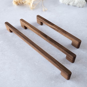 Puede incluir: Tres tiradores de madera para armarios con un acabado marrón oscuro. Cada tirador tiene una barra rectangular con extremos redondeados y una pequeña base rectangular. Disposición diagonal.