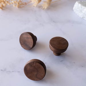 May include: Three round, dark brown wooden cabinet knobs with a flat top and a rounded base are arranged on a white marble surface. The knobs are made of a rich, warm-toned wood, and are likely for furniture.
