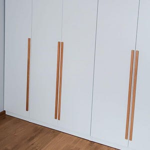 Peut inclure: Une grande armoire blanche avec des poignées en bois et une suspension en osier. L'armoire comporte plusieurs portes et tiroirs. La suspension est en bois tressé et est suspendue au plafond. La pièce a un parquet.