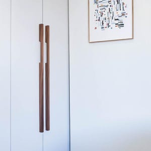 May include: A white wardrobe with two vertical, dark wood handles. A framed abstract art print hangs on the adjacent wall. The artwork features a geometric design with black, red, and blue shapes.