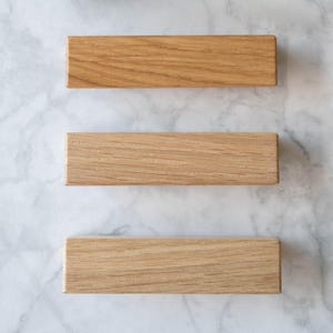 Könnte beinhalten: Drei Holzblöcke in verschiedenen Brauntönen, vertikal auf einer weißen Marmorfläche angeordnet. Die Blöcke bestehen aus Naturholz mit sichtbaren Maserungen.