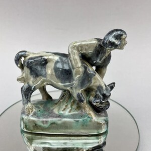 May include: A ceramic figurine of a person riding a black and white spotted cow. The figurine is glazed in shades of green, blue, and white.