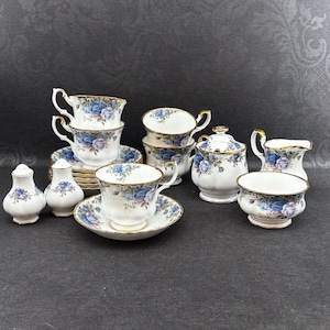 Könnte beinhalten: Ein Vintage-Teeservice mit weißen Porzellantassen, Untertassen und Servierteilen. Das Set ist mit blauen Blumenmustern und goldenen Verzierungen verziert. Enthält Tassen, Untertassen, eine Zuckerdose, einen Sahnekännchen sowie Salz- und Pfefferstreuer.