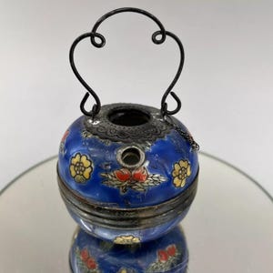 May include: A small blue and white porcelain pot with a black metal handle and a floral design. The pot has a small hole in the top and a chain attached to the handle.