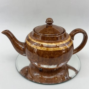 May include: A brown ceramic teapot with a lid and a white stripe around the middle. The teapot is sitting on a mirrored surface.