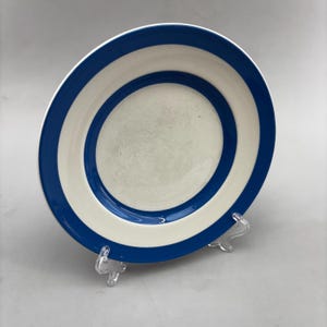 May include: A white ceramic plate with a blue stripe around the rim and a blue band in the center. The plate is displayed on a clear plastic stand. The plate has a simple, clean design.