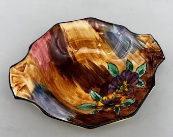 May include: A decorative ceramic dish with a scalloped edge. The interior features a brown, orange, and purple abstract design. The dish is adorned with painted purple and yellow pansies with green leaves, and has a black rim.
