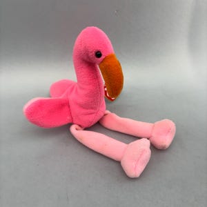 Op de afbeelding: Een flamingo knuffel van felroze stof met een getextureerde oranje snavel en zwarte plastic ogen. Het knuffeldier heeft lange, slanke poten en een klein rood hartvormig label bevestigd aan de zijkant van de kop bij de snavel.