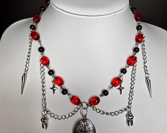 May include: A gothic-style necklace featuring red and black beads, silver chains, and decorative charms. The necklace includes a locket, crosses, and dangling spikes and teeth. The necklace is displayed on a white mannequin bust.