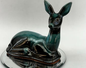 May include: A ceramic deer figurine with a glossy green and black glaze. The deer is lying down with its head turned to the side. The figurine is sitting on a mirrored base.