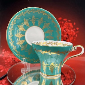 May include: A teal teacup and saucer set with gold leaf design. The teacup has a curved handle and a white interior. The saucer and teacup are decorated with gold leaf patterns. The set is displayed on a reflective surface.