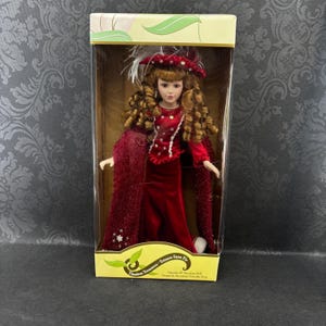 May include: A porcelain doll in a box. The doll is dressed in a red velvet gown, cape, and a hat with feathers. The doll has long, curly brown hair. The box is labelled "Timeless Treasures".
