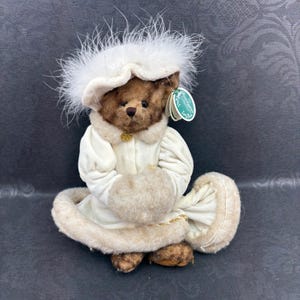Op de afbeelding: Een bruine pluche teddybeer draagt een crèmekleurige fluwelen jas met imitatiebont en een bijpassende hoed versierd met witte veren. De beer heeft een donkerbruine neus en een groen label aan het oor met de tekst The Remington Collection.
