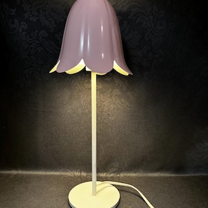 May include: A purple table lamp with a white base and a bell-shaped shade. The lamp is turned on and the light is shining.