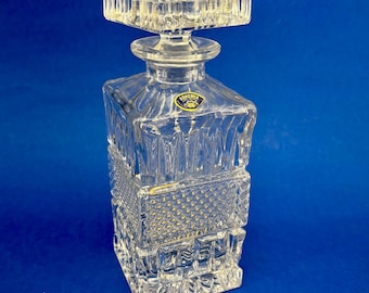 May include: A clear, square-shaped crystal decanter with a matching stopper. The decanter features a geometric design with vertical and diamond-shaped cuts. A small, round sticker with the word "Bohemia" is visible on the front.