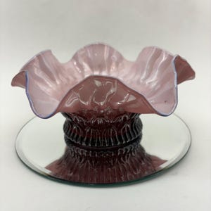 Könnte beinhalten: Eine gerüschte rosa Glasschale mit einem gewellten Rand und einem strukturierten Boden. Die Schale steht auf einer spiegelnden Oberfläche.