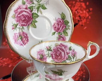 May include: A white teacup and saucer set with a pink rose floral design and gold trim. The teacup has a curved handle and sits on a small, round base. The saucer has a scalloped edge. The set is displayed on a reflective surface.
