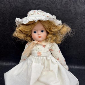 May include: A porcelain doll with blonde curly hair, blue eyes, and rosy cheeks. The doll wears a white dress with a floral pattern and a matching bonnet. A peach-colored ribbon is tied around the neck. The doll is a vintage collectible.