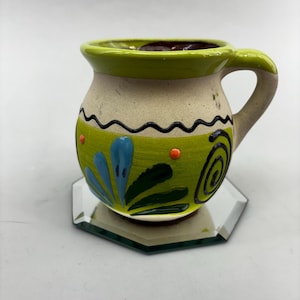 Op de afbeelding: Een groene, witte en blauwe keramische mok met een bloemenmotief. De mok heeft een rond lichaam en een handvat. Hij staat op een gespiegelde achtkantige onderzetter.