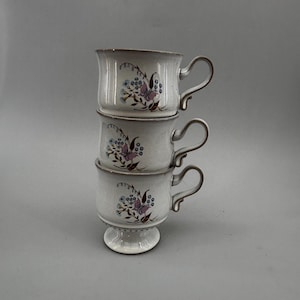 May include: Three stacked white ceramic teacups with a floral design featuring purple butterflies and blue flowers. Each cup has a curved handle and a pedestal base on the bottom cup. The rims are a darker brown color.
