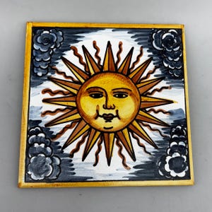 Könnte beinhalten: Quadratische Keramikfliese mit einer lächelnden Sonne mit gelben und braunen Strahlen. Die Sonne ist vor einem weißen Hintergrund mit dunkelgrauen Wolken in jeder Ecke platziert. Die Fliese hat einen goldenen Rand.
