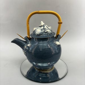 Peut inclure: Une théière en céramique bleue avec un motif de coquillages et une poignée en bambou. La théière a un couvercle avec un motif de coquillages et deux bâtonnets de bambou qui sortent du bec.