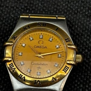May include: A gold and silver two-tone Omega Constellation wristwatch with a gold face and diamond hour markers. The watch face also features the Omega logo and the word "Constellation".