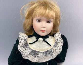 Könnte beinhalten: Eine Porzellanpuppe mit blondem Haar und braunen Augen, bekleidet mit einem dunkelgrünen Samtkleid. Das Kleid hat einen cremefarbenen Spitzenkragen mit einer schwarzen Schleife und einer kleinen weißen Blume am Ausschnitt.