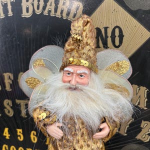 Peut inclure: Une figurine décorative représentant un personnage barbu portant un chapeau texturé doré avec un pompon et des paillettes dorées. Le personnage a une longue barbe blanche, des sourcils blancs et des ailes translucides avec des paillettes dorées.