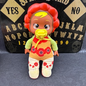 Op de afbeelding: Een pop met een rode bloemvormige hoofdband, geel haar en een gele fopspeen. De pop draagt een outfit met bloemenprint met groene mouwen en een witte broek met rode bloemaccenten. De achtergrond bevat een Ouija-bord.