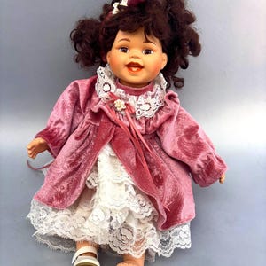 May include: A doll with dark brown curly hair, brown eyes, and an open mouth. The doll wears a pink velvet coat with a white lace collar and a white lace dress underneath. A small white flower accent is attached to the collar with pink ribbons.