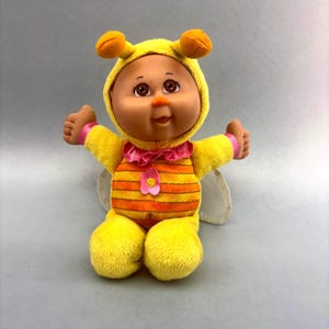 May include: A plush doll dressed in a yellow bee costume with a hood featuring antennae. The doll has a round face, brown eyes, and a striped orange and yellow body with a pink ruffled collar, a pink flower accent, and translucent wings.