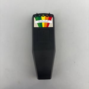 May include: A black handheld electromagnetic field meter featuring a top display with a colour-coded scale ranging from 1.5 to 20+ milliGAUSS (mG). The device includes five small LED indicator lights above the scale and a square battery cover.