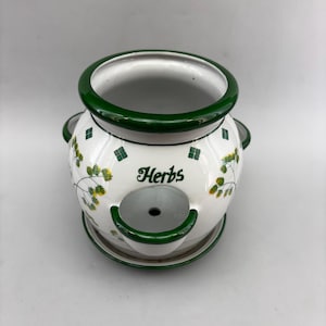 May include: White ceramic herb planter with green trim and the word "Herbs" in a script font. The planter features a round shape with a small opening and a matching saucer. Decorative yellow and green floral accents and small green squares adorn the planter.