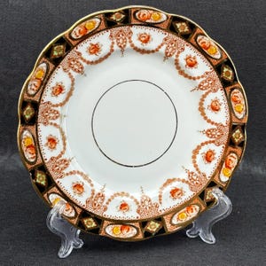 May include: A white porcelain plate with a scalloped edge and a decorative border of gold, black, and red floral patterns. The plate is on a clear plastic stand.