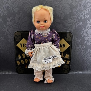 Op de afbeelding: Een vintage pop met blond haar, blauwe ogen en een paarse jurk met witte kanten rand. De pop draagt een crèmekleurig schort en broek. Een label op de schort leest "Chatty Cathy Doll 1961". De pop staat voor een Ouija bord.