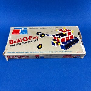 Könnte beinhalten: Vintage Build-O-Fun Master Builder Set Spielzeugkiste. Die Kiste zeigt einen roten, weißen und blauen Spielzeugkran und den Text "Build-O-Fun MASTER BUILDER SET". Die Kiste hat auch das Tupperware Toys Logo.