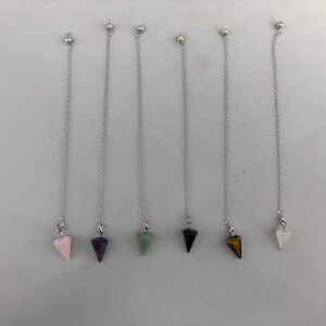 May include: Six pendulums hanging from silver-toned metal chains. Each pendulum features a different gemstone in a pointed, conical shape. The stones include light pink, dark purple, light green, solid black, brown striped, and clear white quartz.