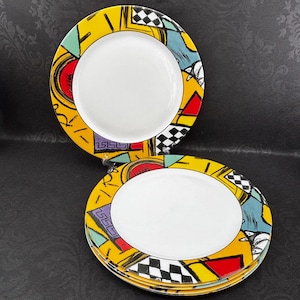 May include: A set of decorative plates with a white center and a colorful border. The border features a yellow background with geometric shapes in red, black, blue, and green. The plates have a unique, artistic design.