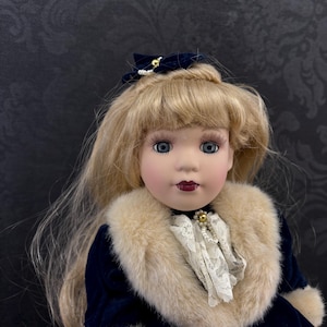 May include: A porcelain doll with long blonde hair, blue eyes, and dark red lips. The doll wears a dark blue velvet coat with a beige faux fur collar, a white lace jabot with a pearl accent, and a matching dark blue hair bow with a gold bead.